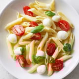 A close-up of Caprese Salad Pasta tossed in olive oil with glistening mozzarella pearls and vibrant cherry tomato halves, garnished with fresh basil.  
