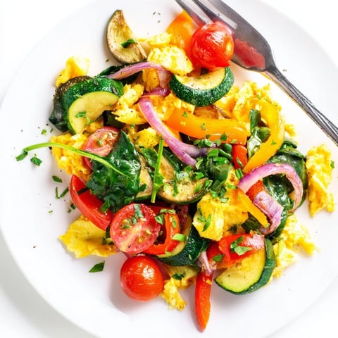 Fluffy scrambled eggs with vibrant red bell pepper, spinach, and cherry tomatoes, garnished with fresh herbs on a white plate.  
