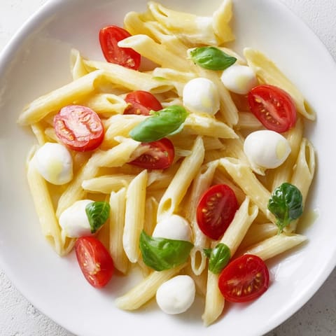 A close-up of Caprese Salad Pasta tossed in olive oil with glistening mozzarella pearls and vibrant cherry tomato halves, garnished with fresh basil.  